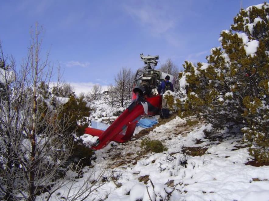 Rear view of the crashed Eurocopter AS350 B3 helicopter positioned on a snow-covered slope among dense shrubs and small trees. The tail boom extends downhill, and the main rotor system is visibly damaged above the cabin. A person in winter clothing stands near the cockpit area as snow blankets the surrounding terrain.