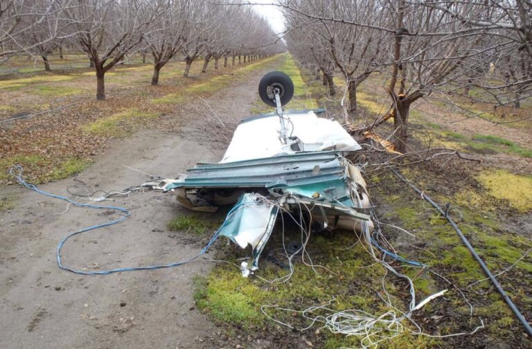 Wreckage of the Piper PA-32 in an orchard near the crash site. A section of the fuselage rests inverted with the landing gear pointing upward, surrounded by torn wiring and broken tree limbs. NTSB photo.