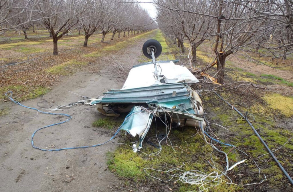 Wreckage of the Piper PA-32 in an orchard near the crash site. A section of the fuselage rests inverted with the landing gear pointing upward, surrounded by torn wiring and broken tree limbs. NTSB photo.
