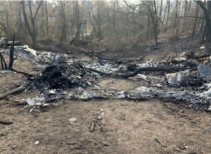 Burned aircraft wreckage scattered across a wooded crash site, with charred debris and scorched ground visible among the trees.