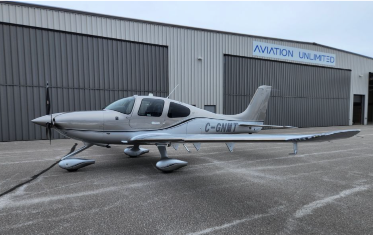 Silver single-engine Cirrus aircraft parked on a ramp in front of large hangar doors at Aviation Unlimited, with its Canadian registration visible on the fuselage.