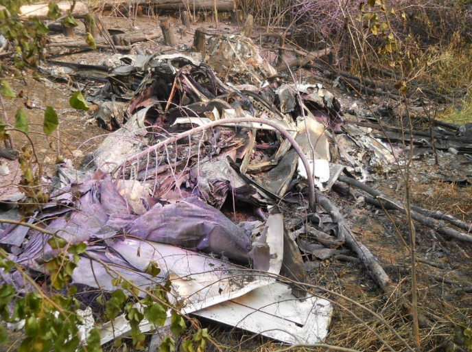 Burned and heavily fragmented aircraft wreckage lying in a wooded area, with scorched debris, damaged metal panels, and dry brush surrounding the crash site.