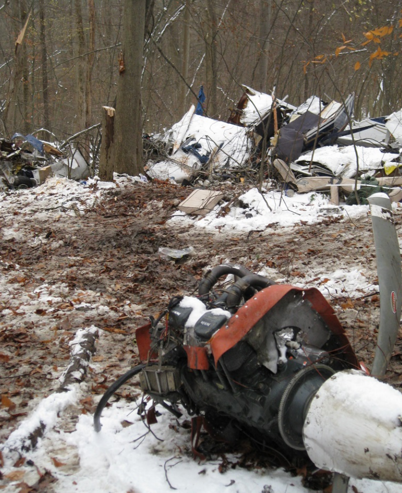 Aircraft wreckage scattered in a snowy wooded area, with a damaged engine section in the foreground and fuselage debris among the trees.