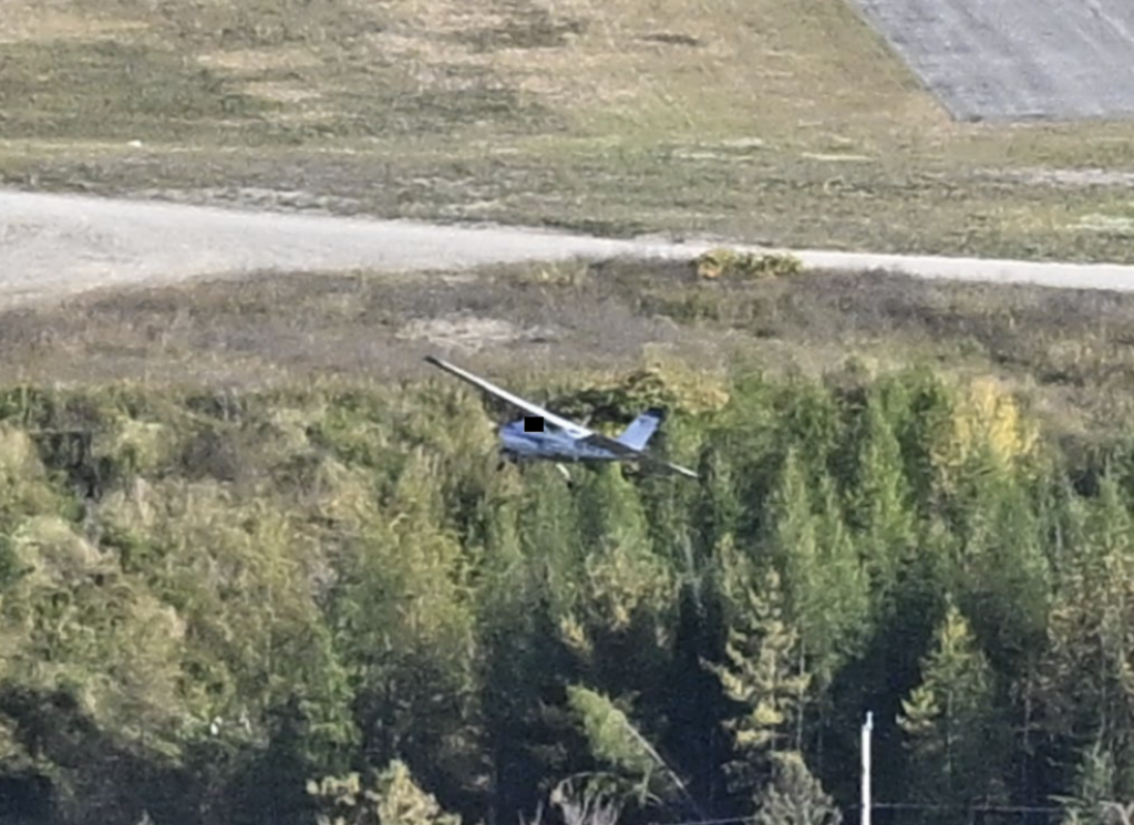 Small airplane flying low over trees and brush near an airport or cleared field.