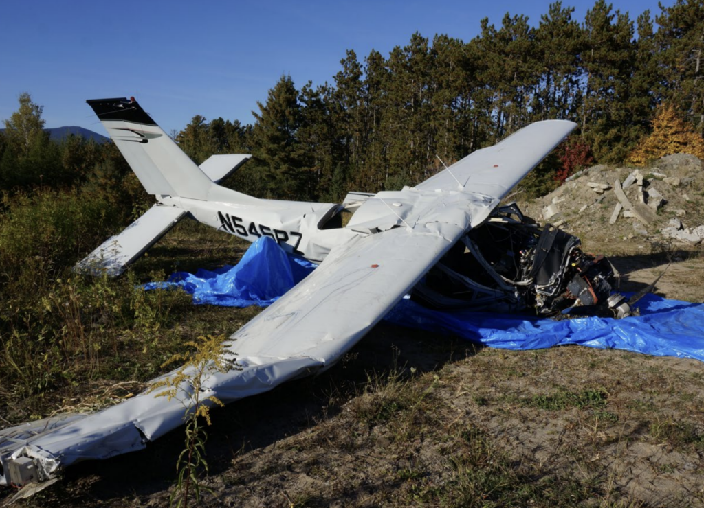 Damaged white airplane wreckage resting on a blue tarp in a wooded area, with the nose crushed and one wing bent.