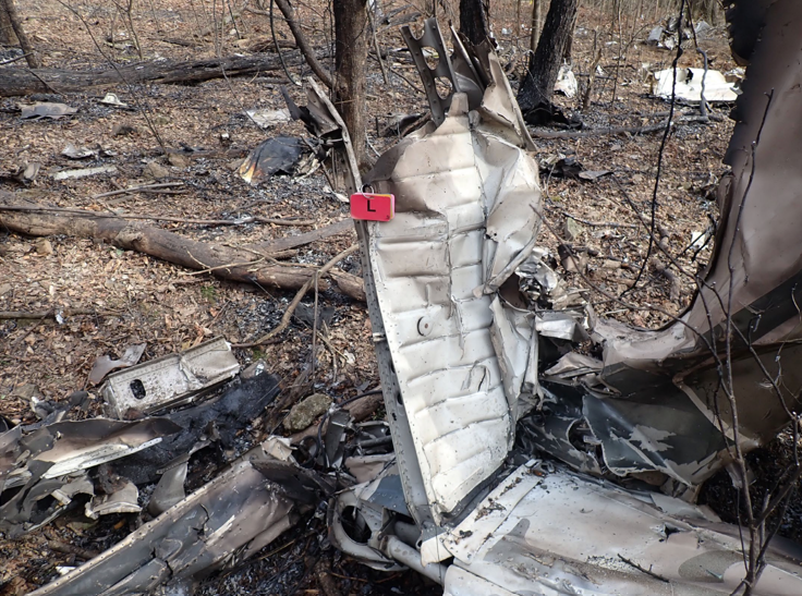 Burned aircraft wreckage scattered in a wooded area, with a torn vertical tail section and debris among trees and charred ground.