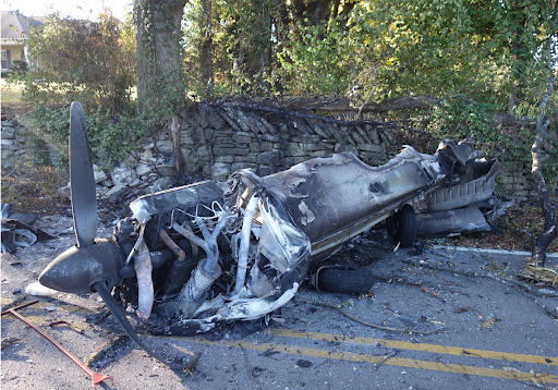 Burned aircraft wreckage on a road beside a stone wall, with the propeller and charred fuselage visible.