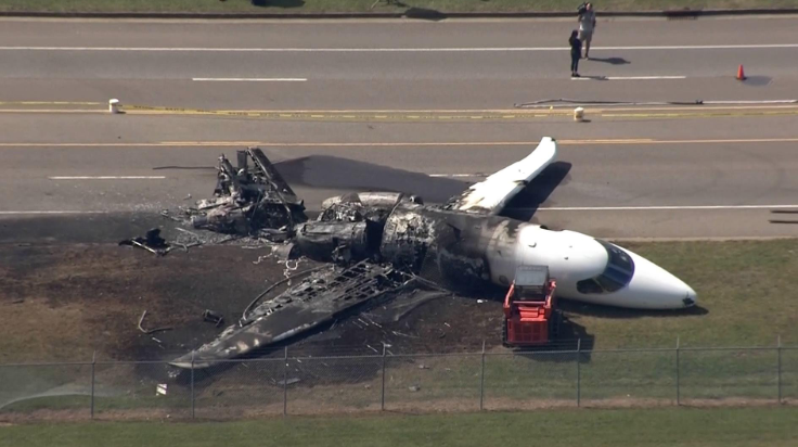 Aerial view of burned business jet wreckage beside a road and fence after a runway excursion, with the nose section largely intact and the fuselage heavily fire-damaged.