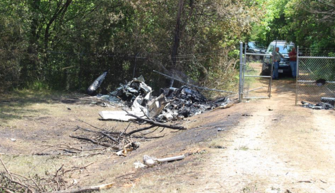 Burned aircraft wreckage scattered beside a dirt road and chain-link gate in a wooded area.