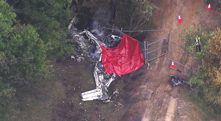 Aerial view of aircraft wreckage near trees and a fence, with part of the debris covered by a red tarp and traffic cones marking the scene.