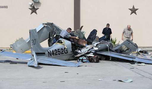Crashed Cirrus SR20 wreckage near a building, with the fuselage broken apart and investigators standing nearby.