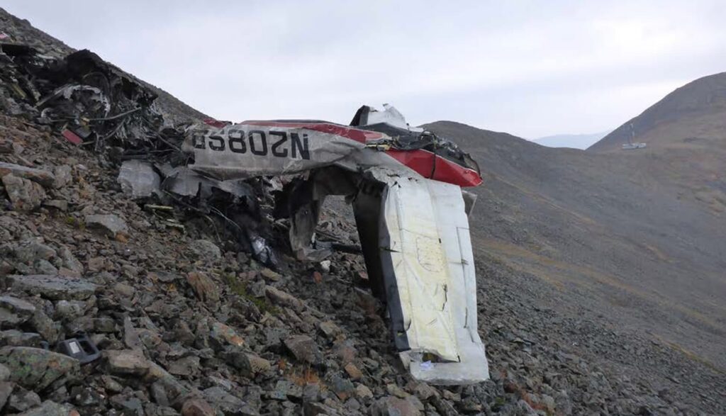 Wreckage of a red-and-white single-engine airplane scattered across a steep, rocky mountainside, with the broken fuselage and wing resting among debris under an overcast sky.