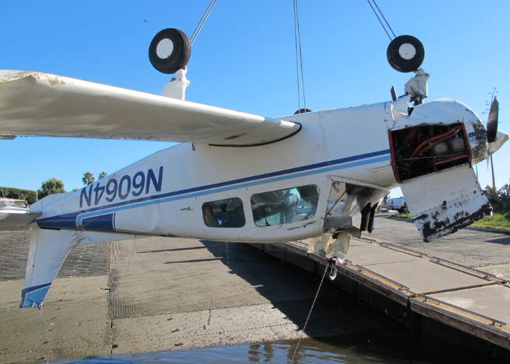 Alt text: Small single-engine airplane, registration N7909N, overturned beside a dock with its landing gear facing upward and visible damage around the nose, engine cowling, and propeller area.