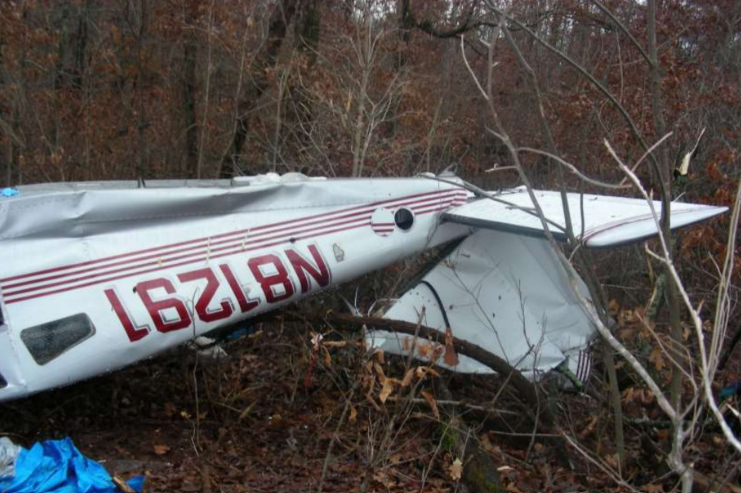 Wreckage of a small white airplane with red striping lying in a wooded area, with the fuselage and tail section visible among branches and fallen leaves.