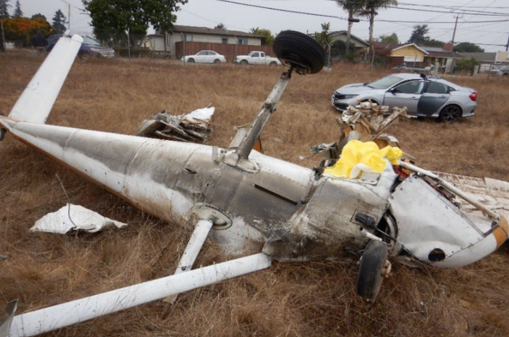 Overturned and heavily damaged small aircraft wreckage lying in dry grass near a road, with broken landing gear visible and a damaged car in the background.