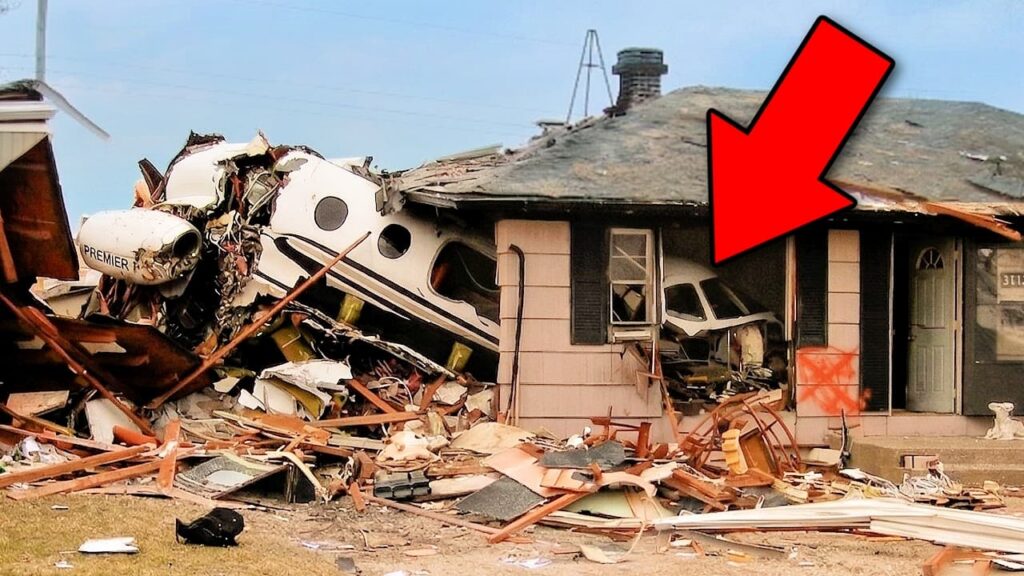A small airplane has crashed into a house, leaving the aircraft and part of the building severely damaged and surrounded by debris. A large red arrow points toward the impact area.