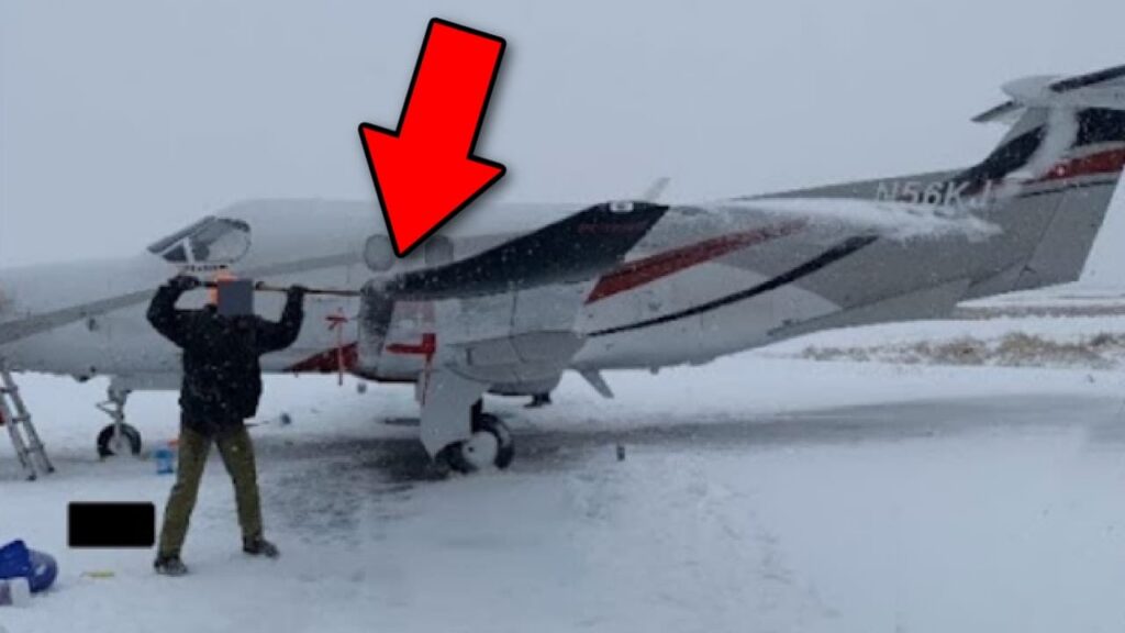 Small propeller airplane on a snow-covered runway during heavy snowfall, with a person standing beside the aircraft near the wing. A large red arrow points toward the airplane’s fuselage and wing area.