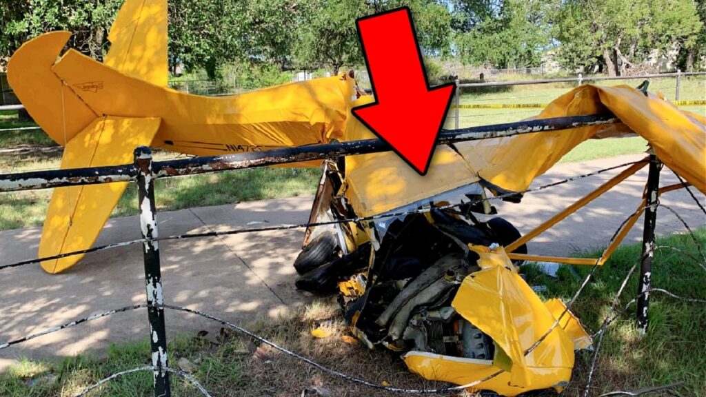 Yellow airplane wreckage crushed against a metal fence in a grassy area, with severe damage to the fuselage and wing. A large red arrow points toward the damaged center section of the aircraft.