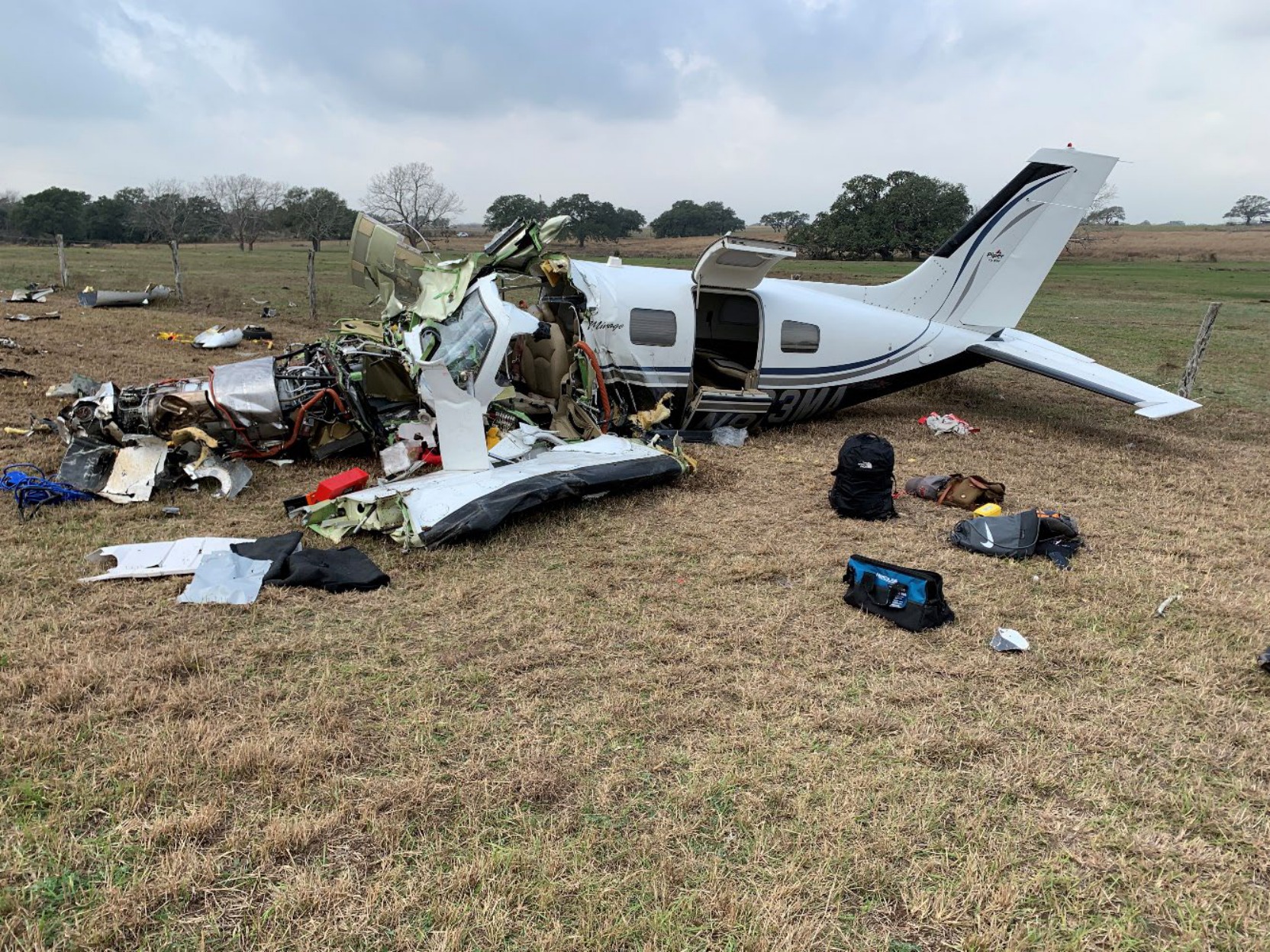 Left side of a wrecked Piper PA-46 in a grassy field, with the cockpit torn open, engine exposed, and cabin door open.