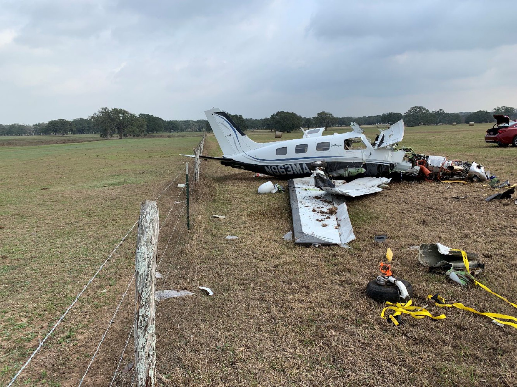 Right side of a wrecked Piper PA-46 beside a wire fence, with a damaged wing in the foreground and the engine exposed.