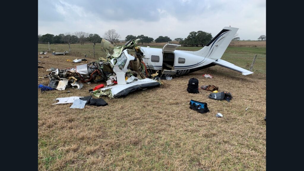 Left side of a wrecked Piper PA-46 in a grassy field, with the cockpit torn open, engine exposed, and cabin door open.