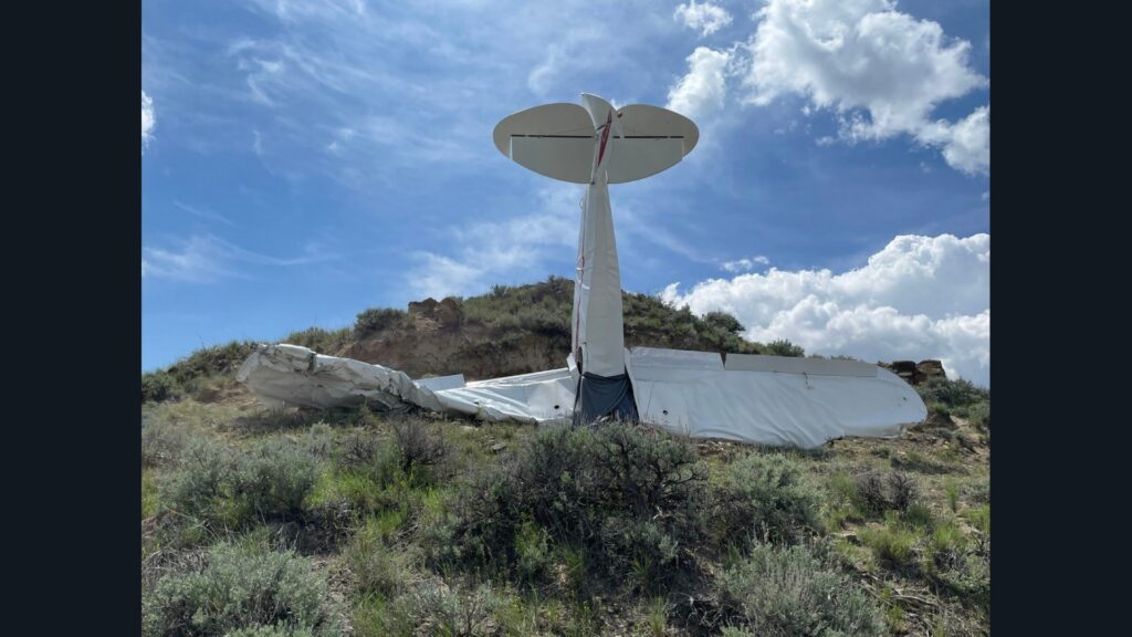 A white aircraft lies inverted or nose-down on a rocky, shrub-covered slope. The tail assembly, including the vertical stabilizer and horizontal stabilizer, is elevated and pointing upward. The fuselage appears crumpled against the terrain. Sparse desert vegetation surrounds the wreckage under a partly cloudy sky.