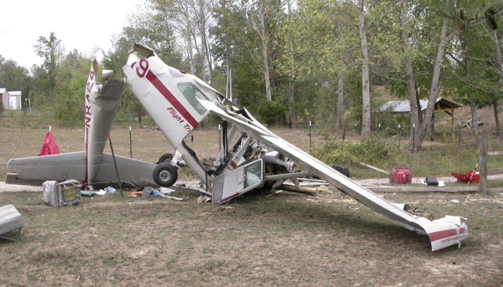 A heavily damaged small white and red propeller aircraft with visible text "Flight Tr..." on its fuselage, lying on a grassy field. The plane is severely crumpled with wings detached and debris scattered around. Trees and a structure are visible in the background.